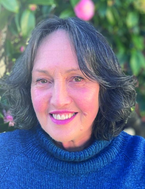Portrait photograph of author Gillian Tompsett, smiling, in a blue jumper in front of flowering trees