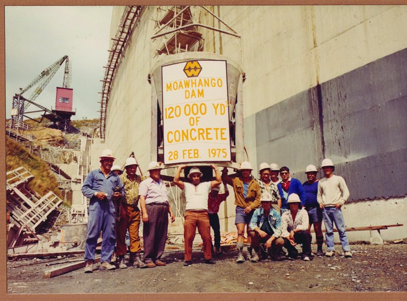 Colour photograph of a Ministry of Works crew in hard hats standing in front of a concrete dam wall, holding up a sign that reads Moawhango Dam, 120,000 yards of concrete, 28 February 1975